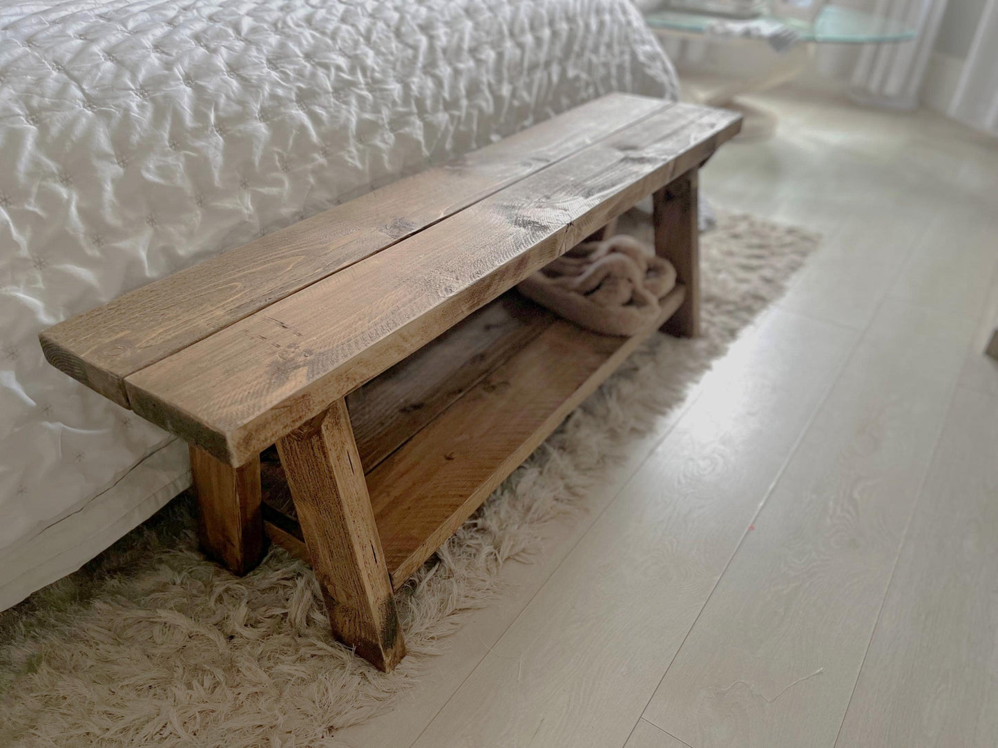 A farmhouse styled, rustic wooden bench with a bottom shelf, in a bedroom setting. (front view)