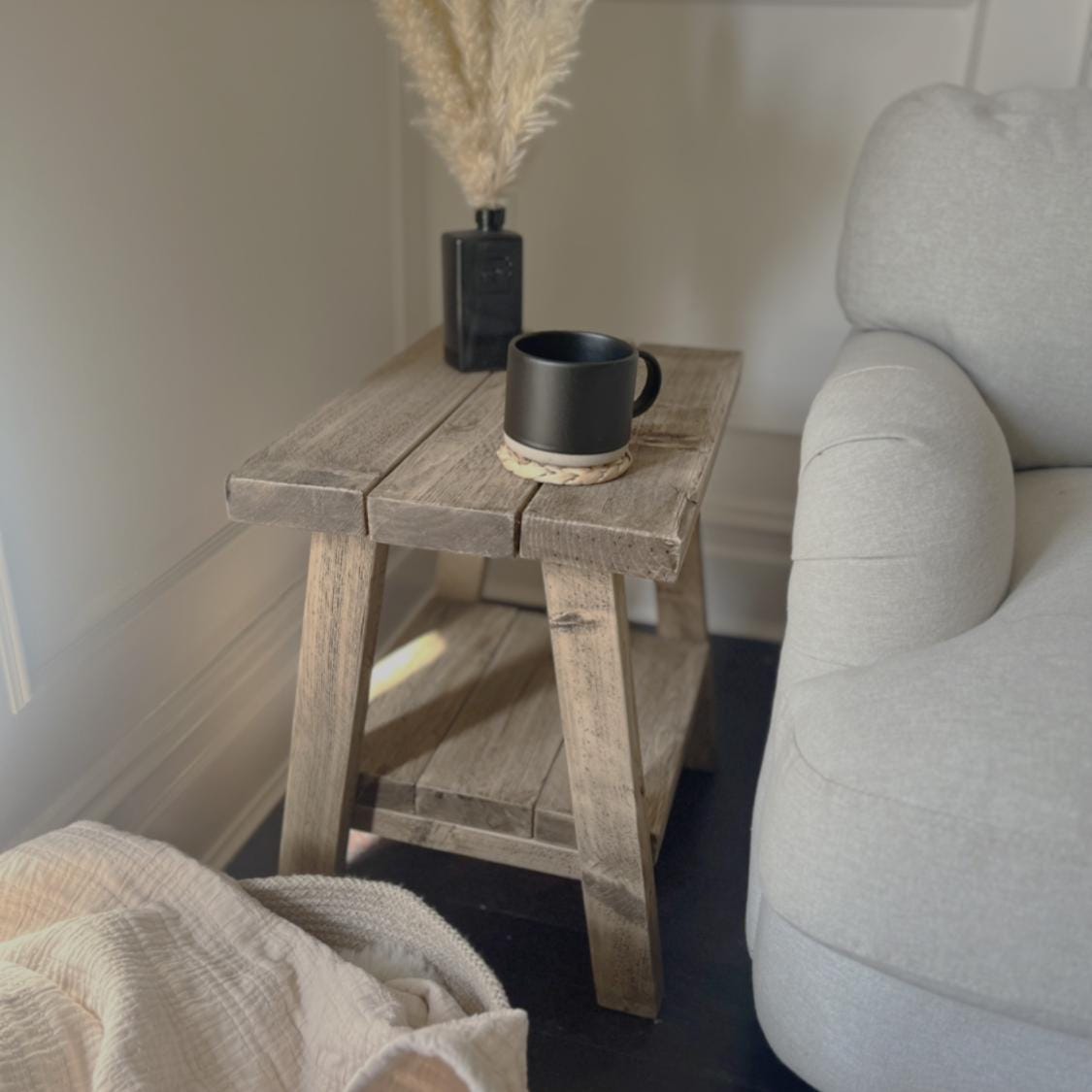 Wooden side table with a black mug and vase next to a gray armchair.