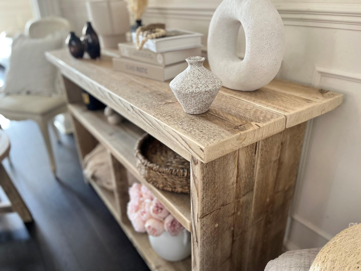 A farmhouse styled, rustic wooden console table with decorative items in a room setting. One upper shelf along the entire length of the table and two lowershelvws at the bottom. Top view in a living room setting