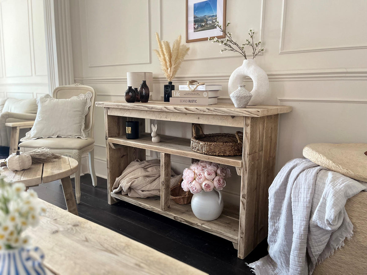 A farmhouse styled, rustic wooden console table with decorative items in a room setting. One upper shelf along the entire length of the table and two lowershelvws at the bottom. Front view in a living room setting