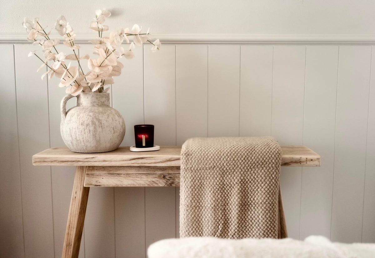 A rustic wooden console table styled with a textured cream ceramic vase holding pale dried flowers, a small dark amber candle, and a beige knit throw blanket draped over the side, set against a light gray paneled wall.