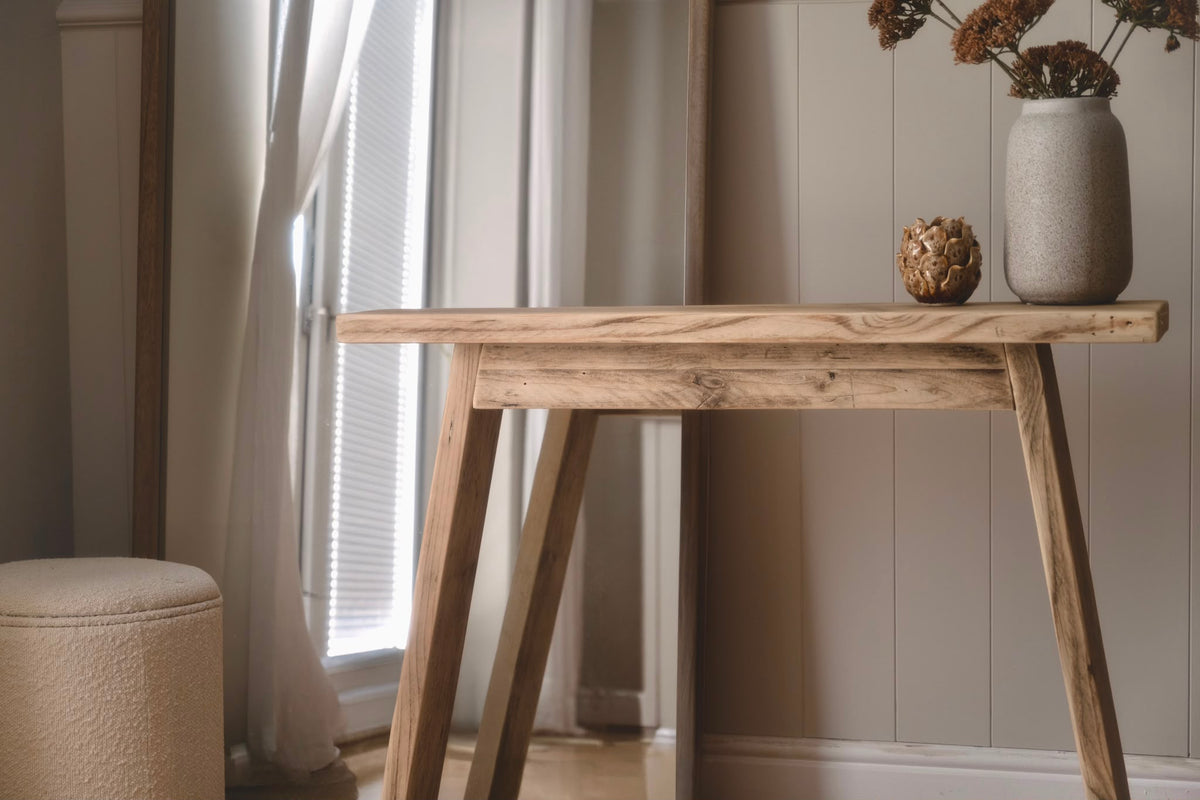 A rustic wooden console table with a thick rectangular top and angled trestle-style legs, styled in a warm interior setting with a vase of dried flowers and a small decorative object on top.
