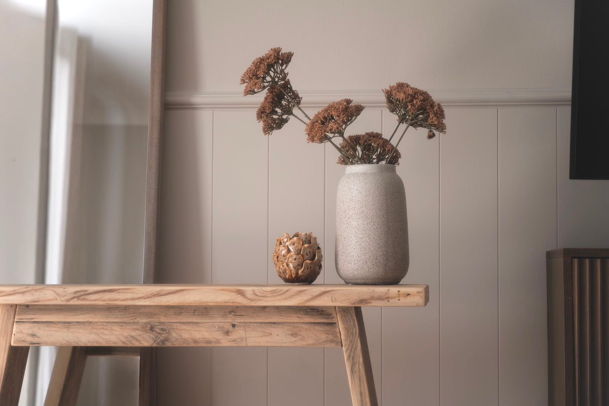A rustic light wood table in a neutral room setting, holding a textured beige ceramic vase filled with dried brown flowers and a small decorative object.