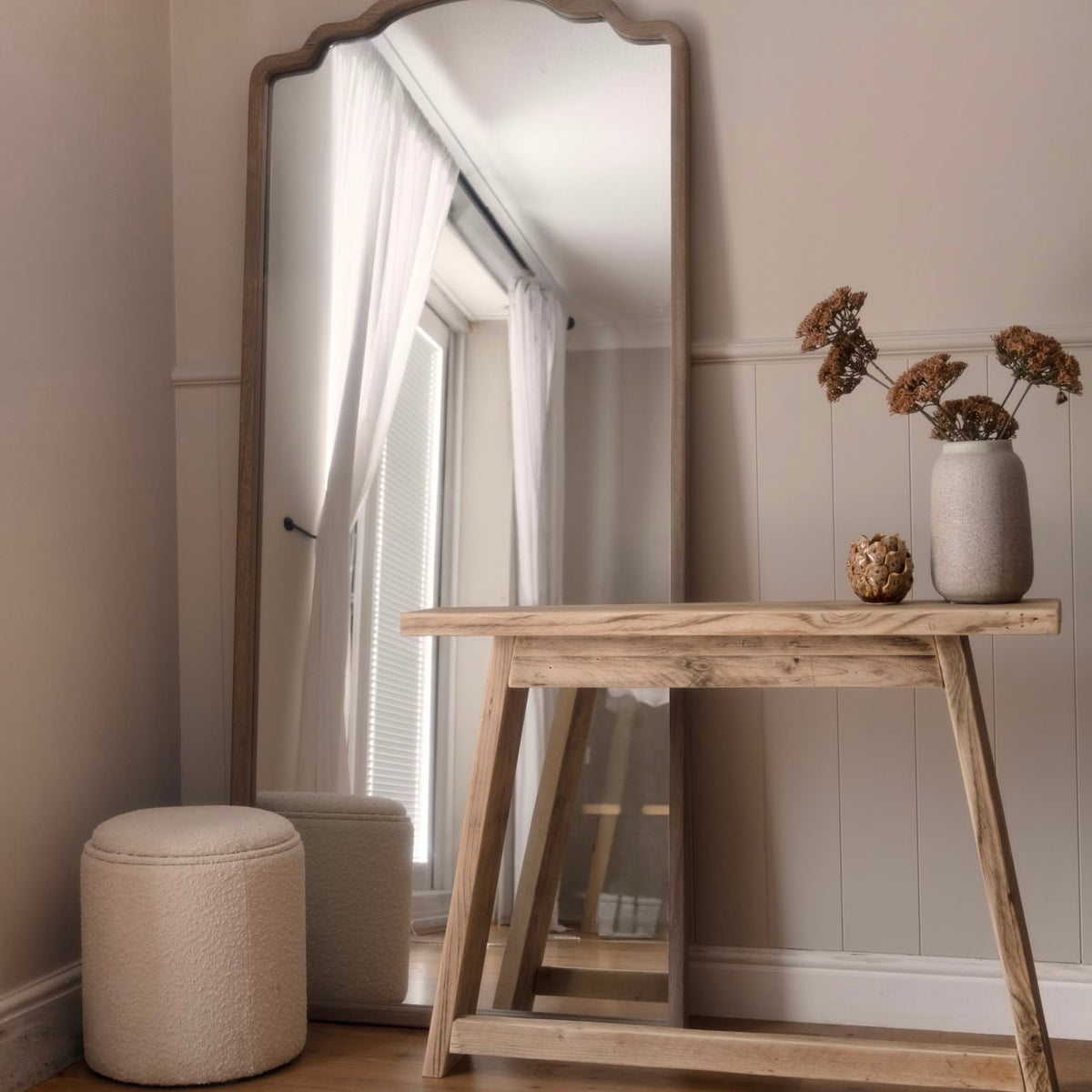 A tall floor mirror with a scalloped top frame stands in a styled room corner next to a rustic wooden console table, with a textured cream pouf and a vase of dried flowers on a light wood floor.