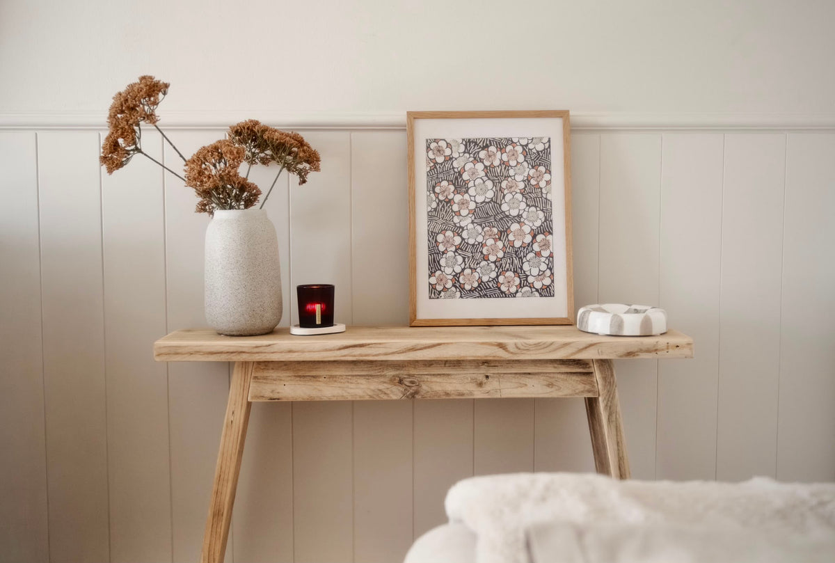 A framed floral art print with a light wood frame is styled on a rustic wooden console table next to a textured vase with dried flowers, a small candle, and a white object, set against a beige paneled wall.
