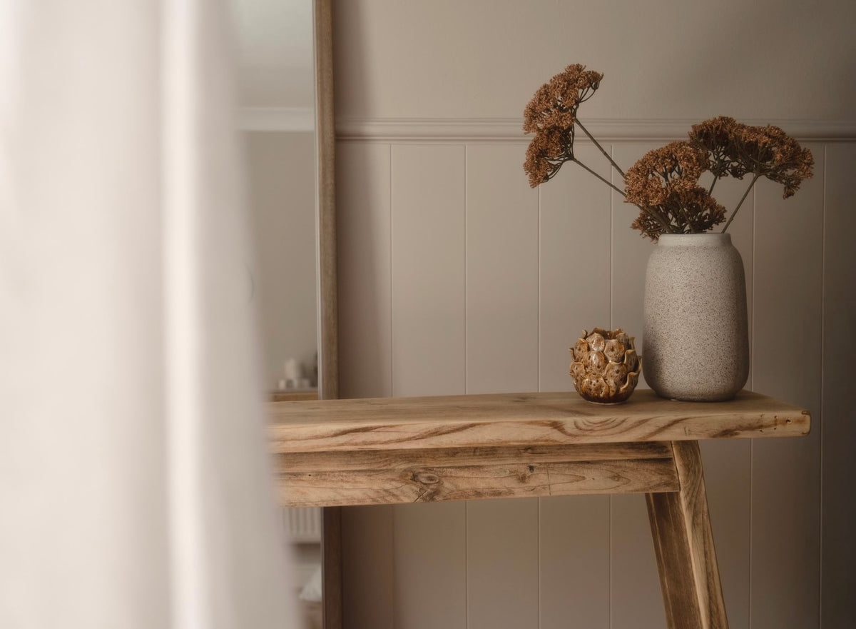 A rustic wooden console table in a warm neutral room, styled with a textured gray ceramic vase holding dried brown flowers and a small decorative sphere.