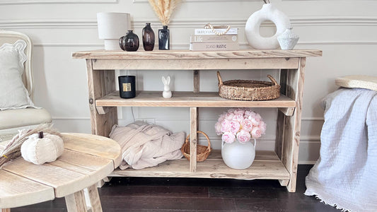A farmhouse styled, rustic wooden console table with decorative items in a room setting. One upper shelf along the entire length of the table and two lowershelvws at the bottom. Front view in a living room setting