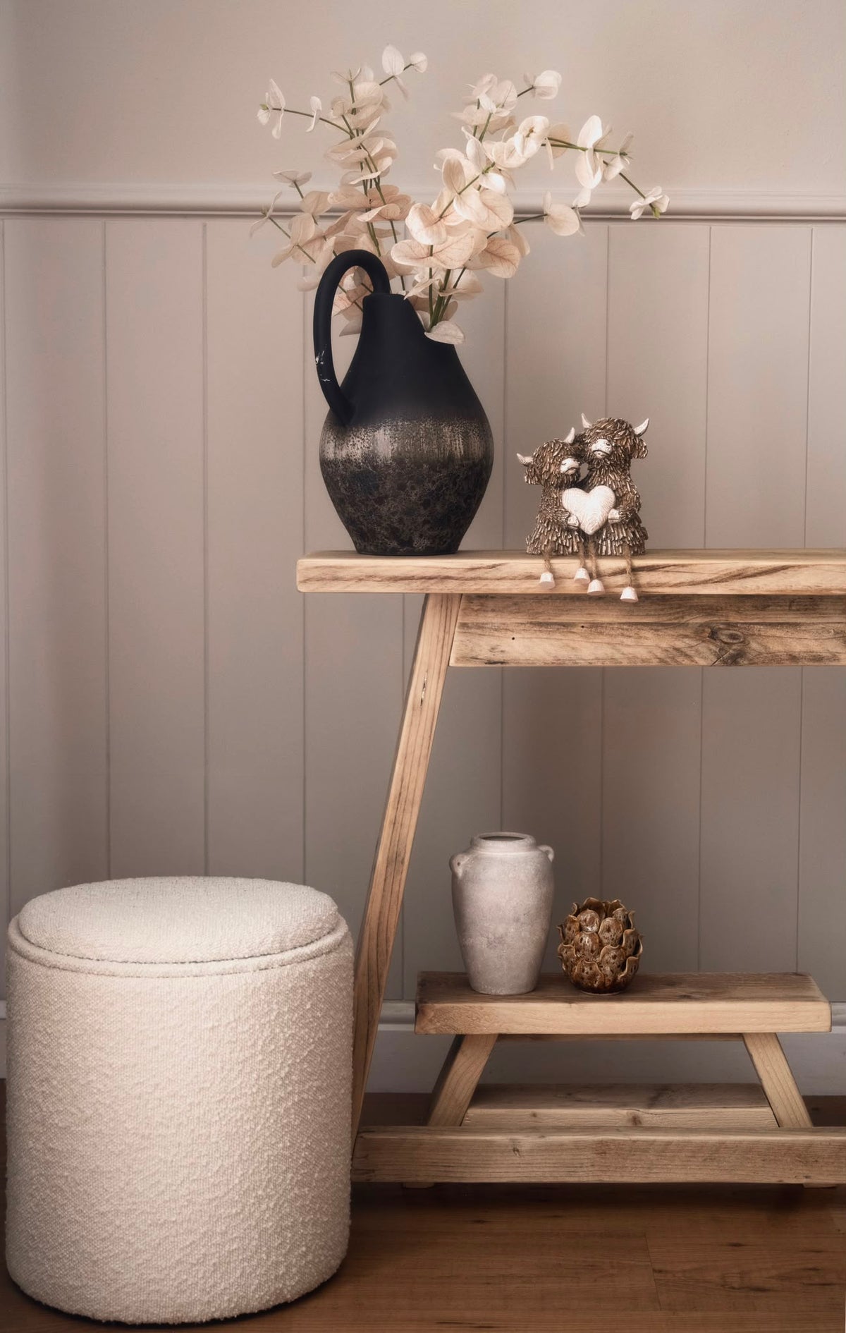 A rustic wooden console table styled in a home interior, featuring a black pitcher vase with dried white flowers, a small owl figurine, and a textured cream pouf in the foreground.