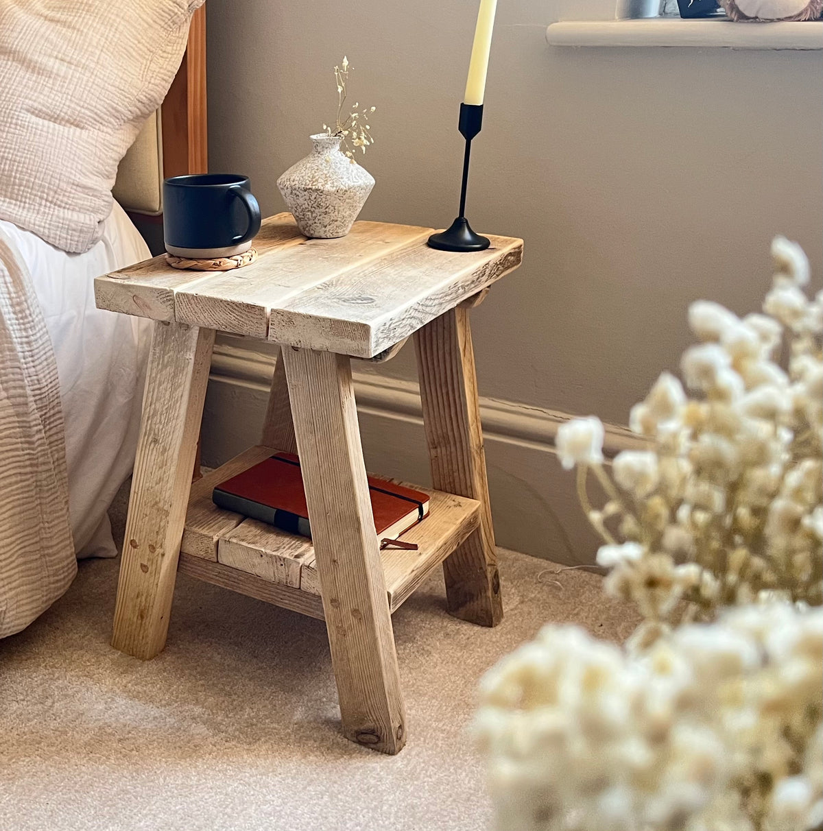 Wooden side table with a cup, vase, and candle next to a bed in a bedroom.