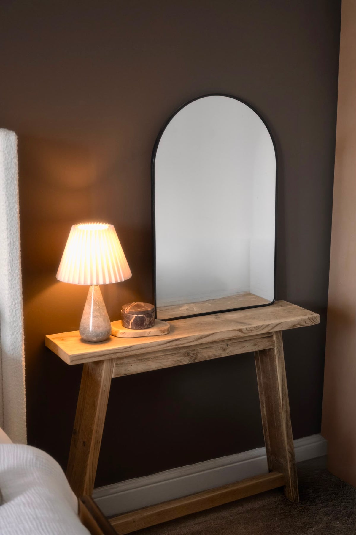 A rustic wooden console table in a bedroom setting, featuring a large arched wall mirror and a lit pleated lamp on top.