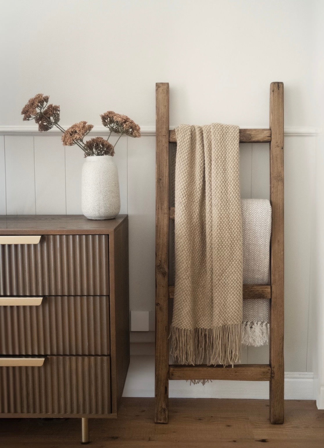 Wooden ladder with a blanket against a white wall next to a wooden dresser.