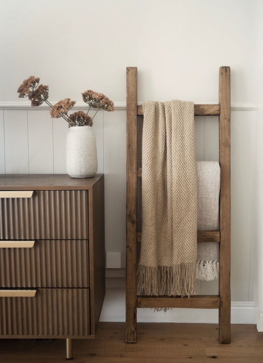 Wooden ladder with a blanket against a white wall next to a wooden dresser.