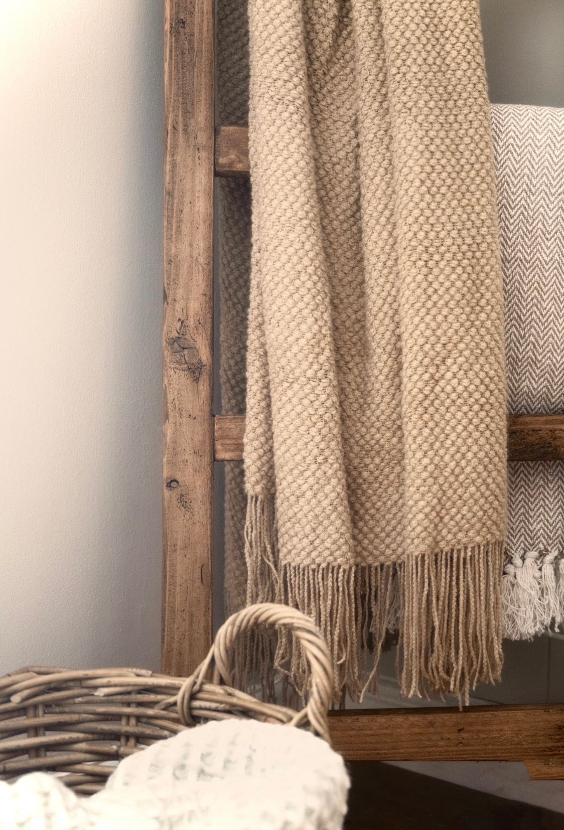 Textured beige blanket with tassels on a wooden shelf against a neutral wall.