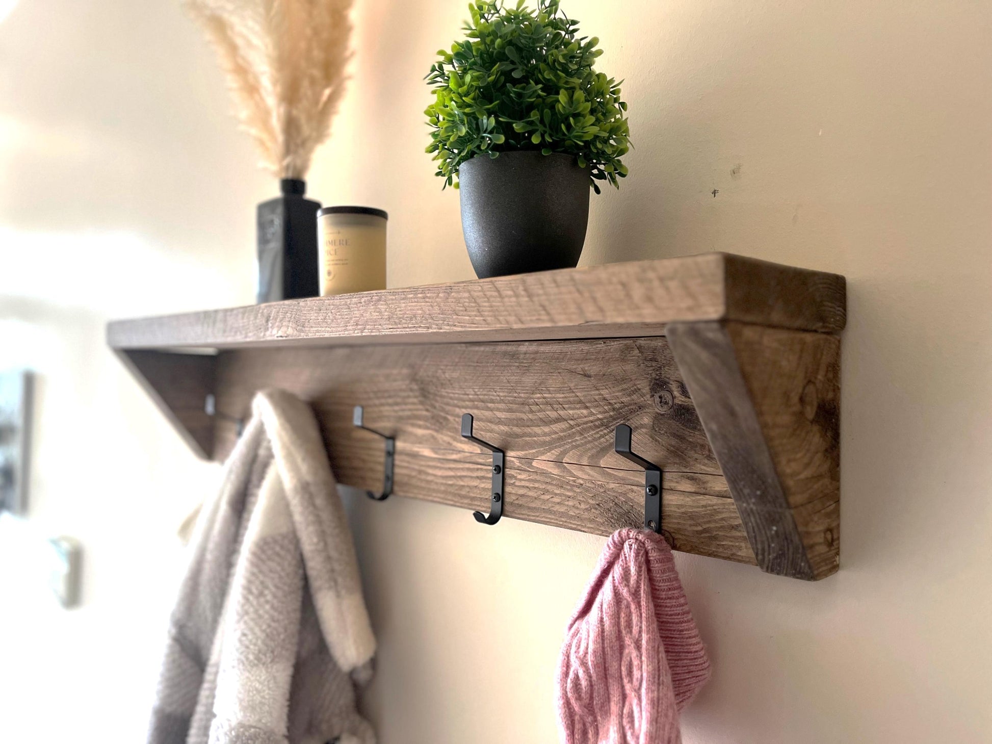 Wooden shelf with hooks holding a towel and pink garment, plants on top against a white wall.
