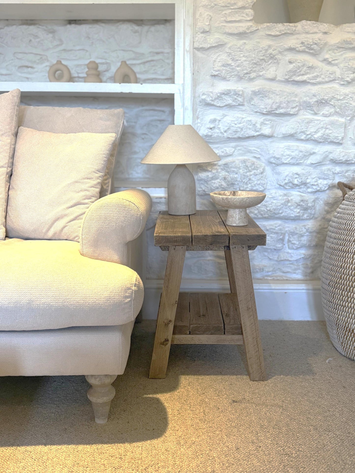 Beige armchair next to a rustic wooden side table with a lamp against a stone wall.