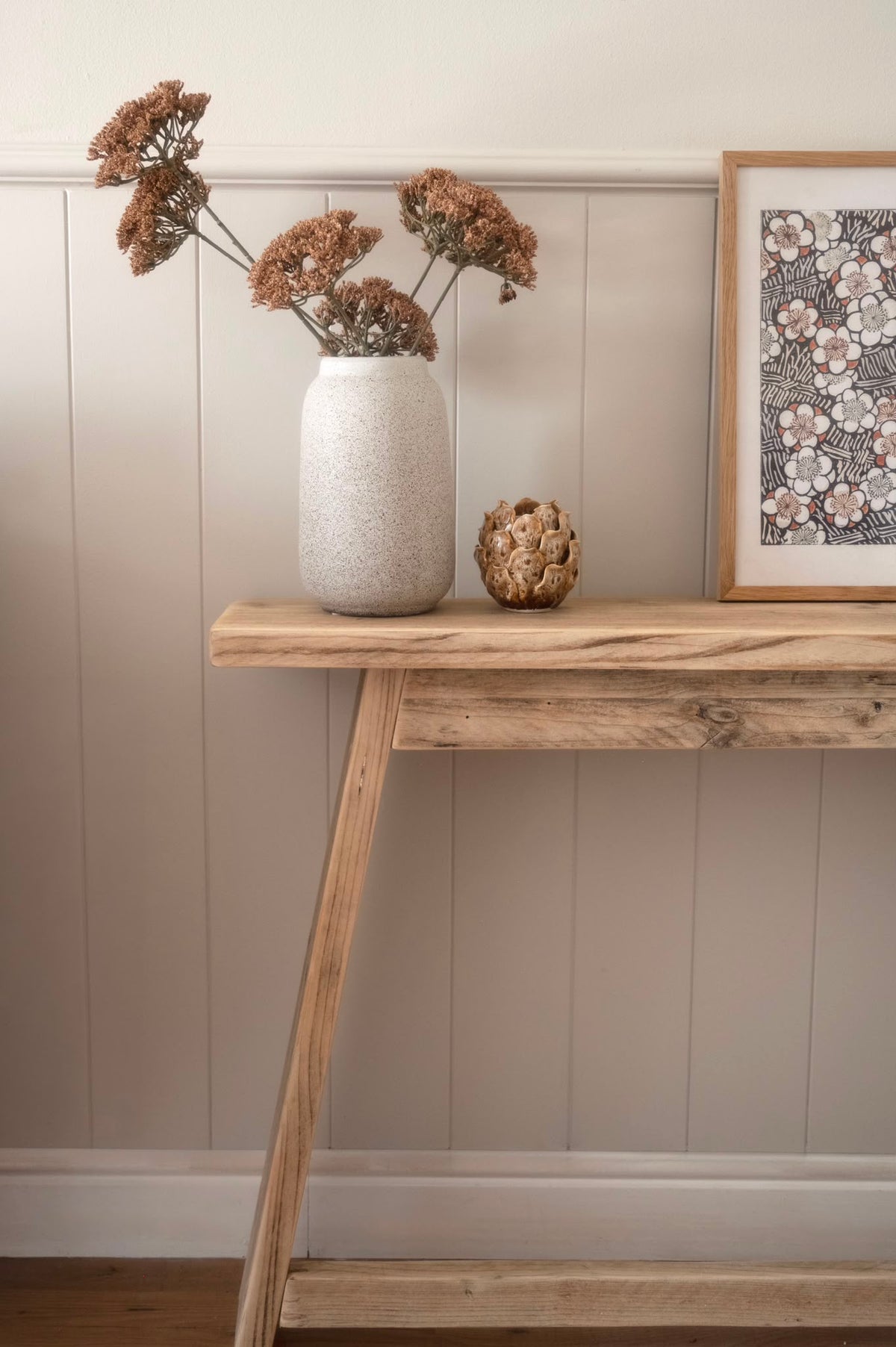 A rustic light wood console table styled with a textured off-white ceramic vase holding dried brown flowers, a small decorative object, and a framed floral print against a beige paneled wall.