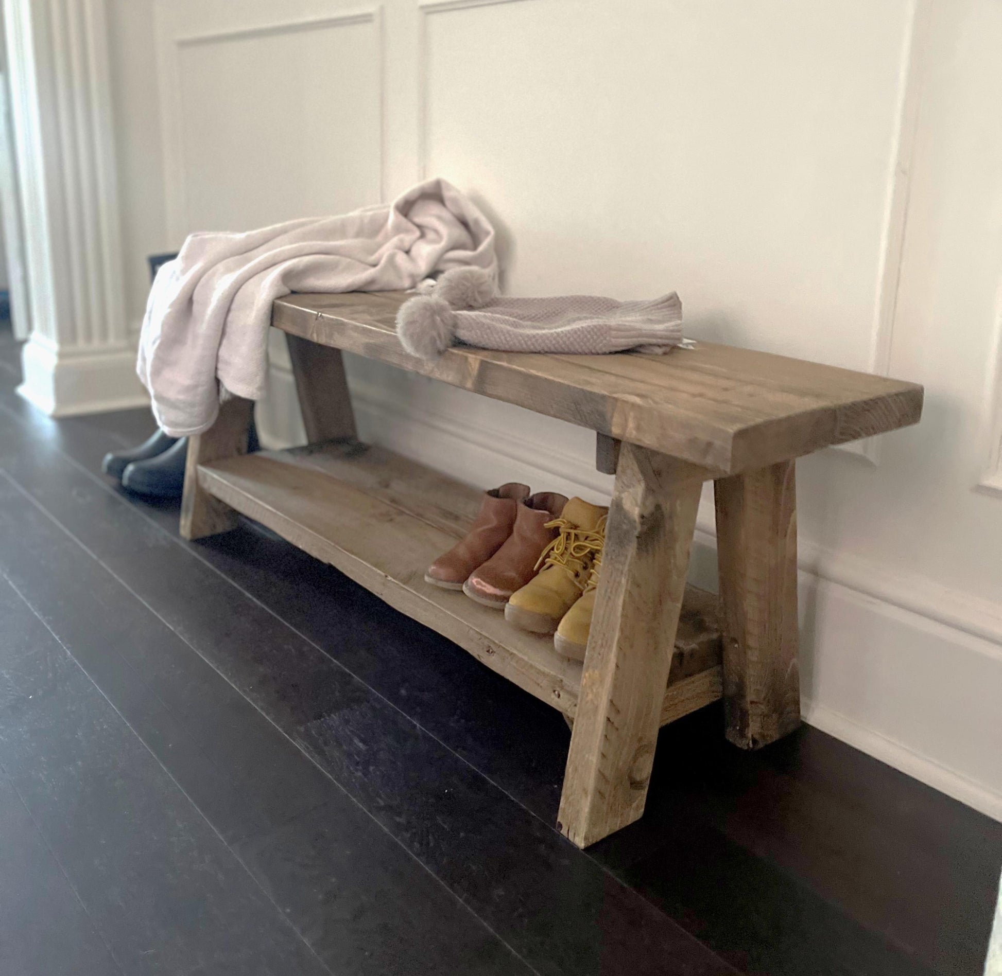 A farmhouse styled, rustic wooden bench with a bottom shelf, in an entryway  setting. (front view)