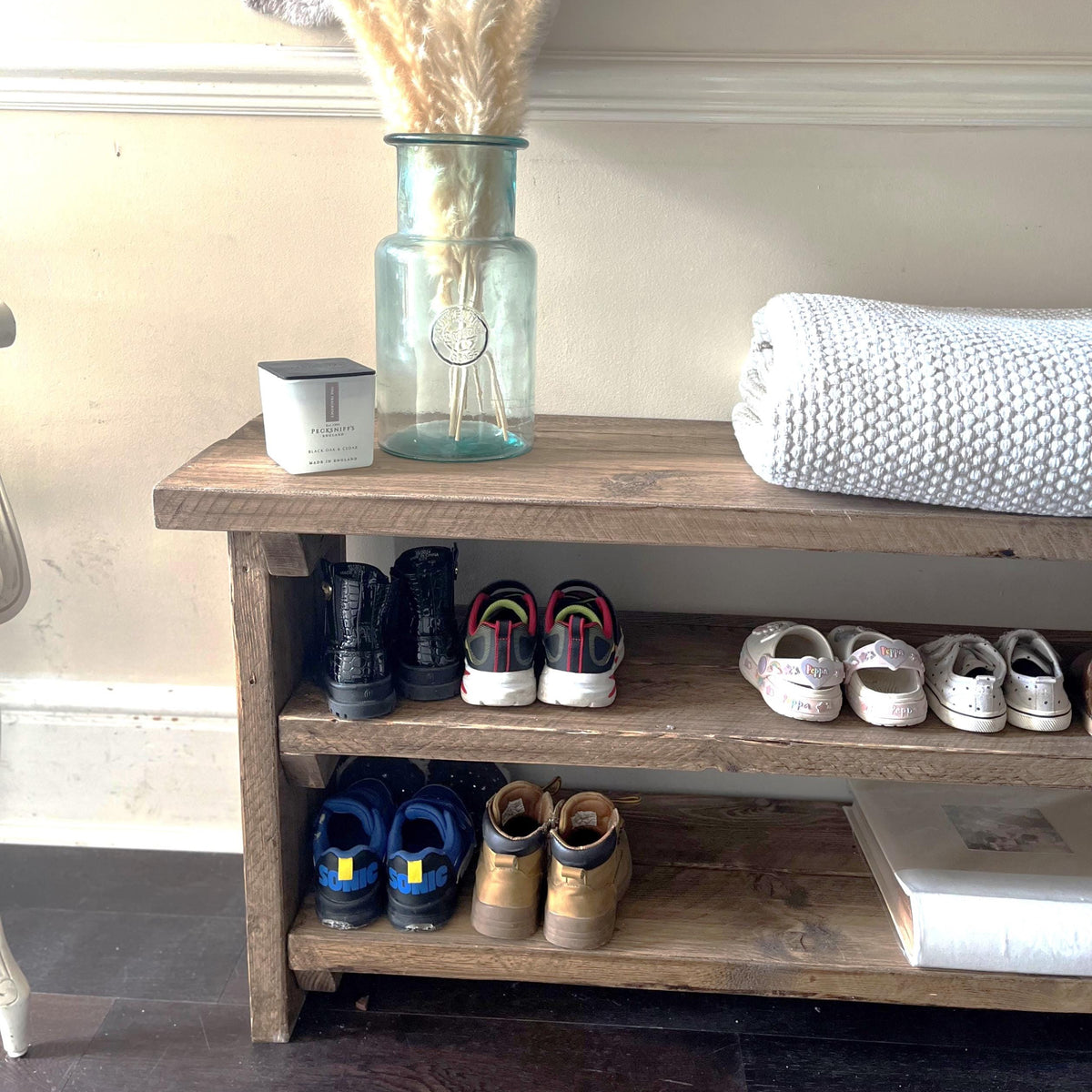 Wooden shoe rack with shoes, a vase with dried plants, and a towel on a light-colored wall background.