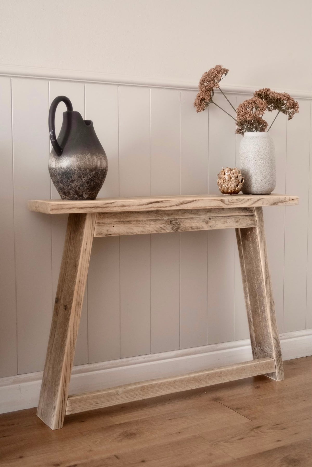A rustic A-frame console table made of light natural wood, styled with a dark textured ceramic pitcher and a white vase with dried flowers against a beige paneled wall and wood floor.